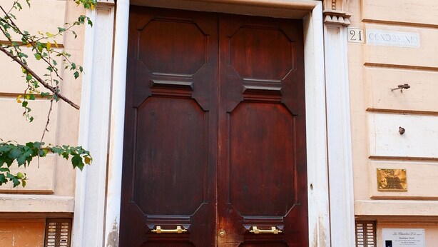 Uma porta dupla de madeira escura e imponente, com a inscrição “A.D. MC MXV” acima.