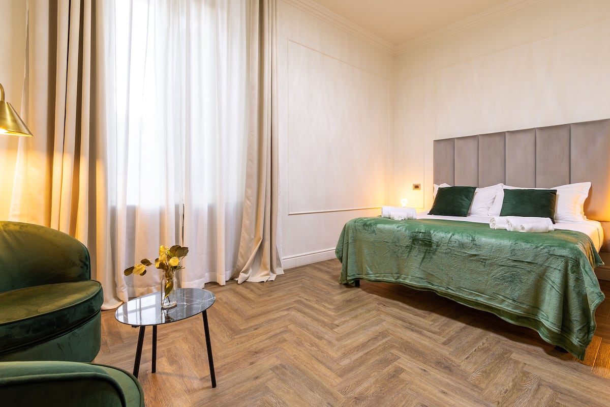 Ein Hotelzimmer mit Bett, grünem Polsterstuhl, einem kleinen Tisch mit Vase und einem Holzboden mit Fischgrätmuster.