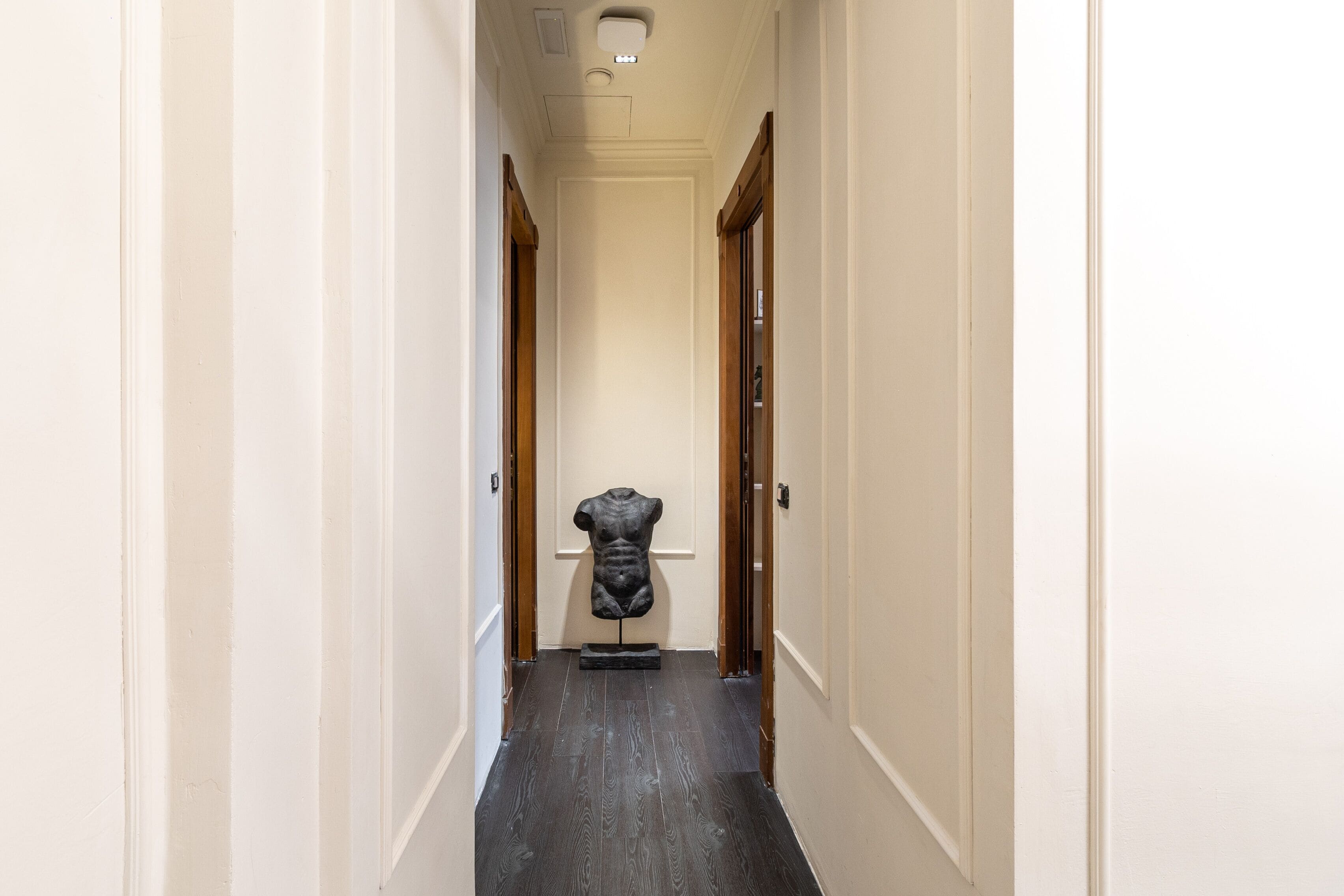 A narrow hallway with a dark wooden floor, white walls, and a sculpture at the end.