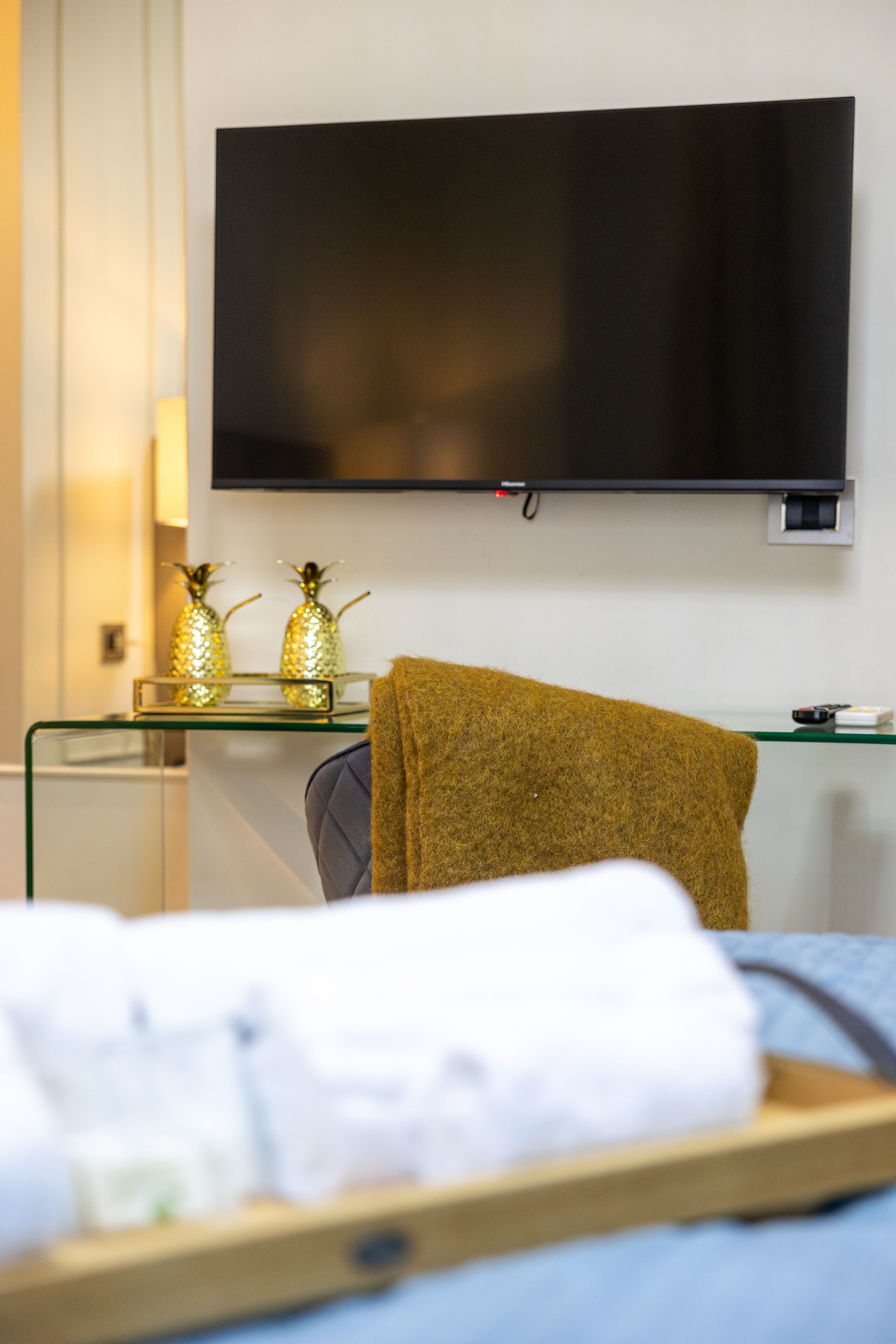 A hotel room with a flat-screen TV mounted on the wall, a glass side table with decorative pineapple-shaped vases, a brown towel on a chair, and a neatly folded white towel on a wooden tray.