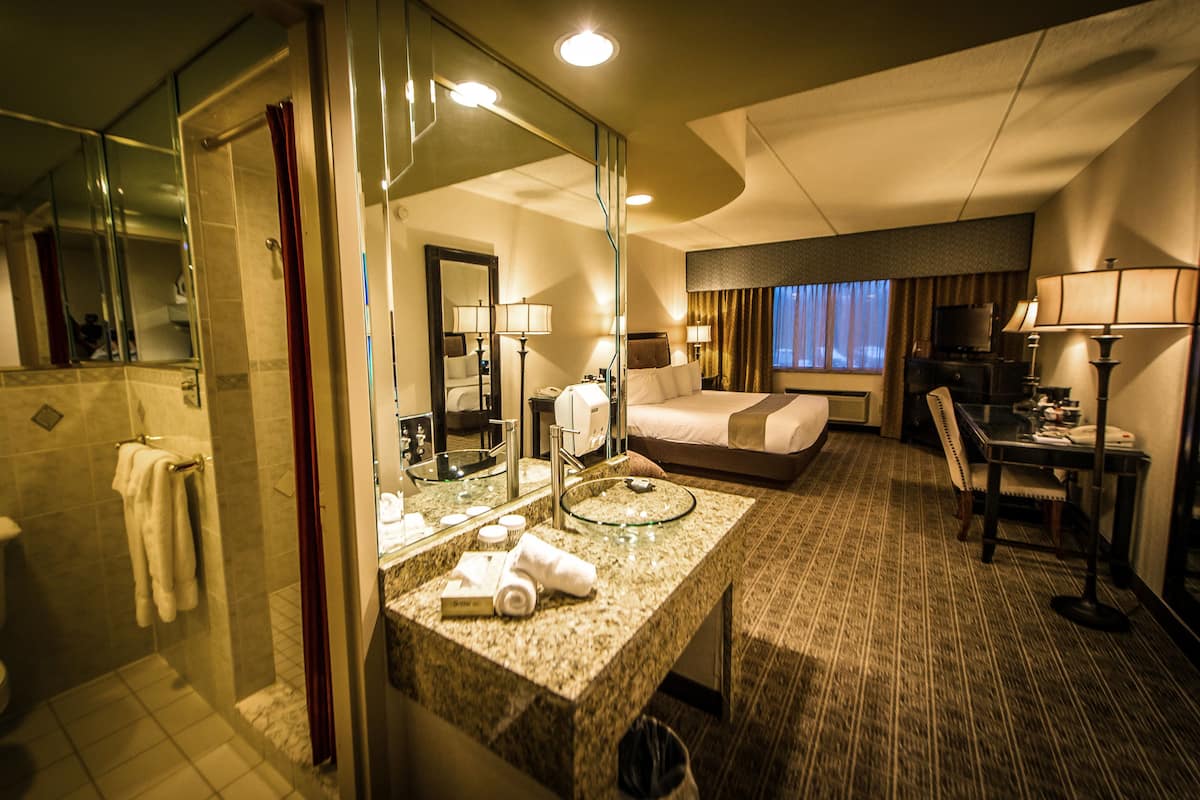 A hotel room with a glass-enclosed shower, a marble bathroom vanity, a bed with a TV, a dining table, and a chair.