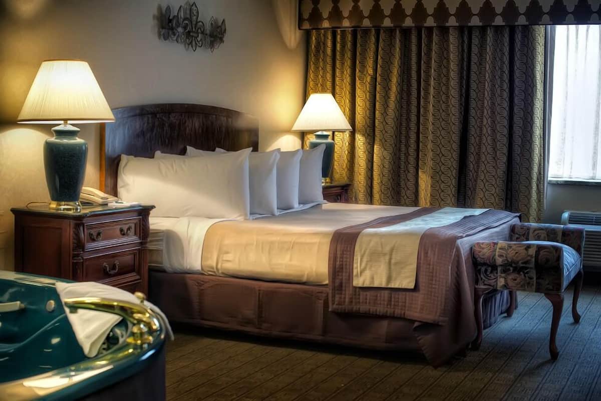 A hotel room with a large bed, two bedside lamps, a wooden nightstand, a green trash bin, and a patterned chair.