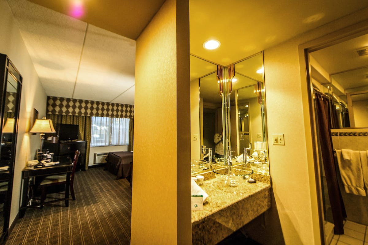 A hotel room with a desk, chair, television, and a large mirror over the sink.