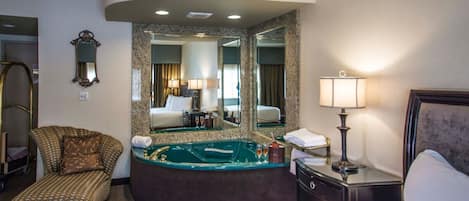 A hotel room with a large bed, a curved seating area, a jacuzzi, a vanity with a mirror, and a lamp.