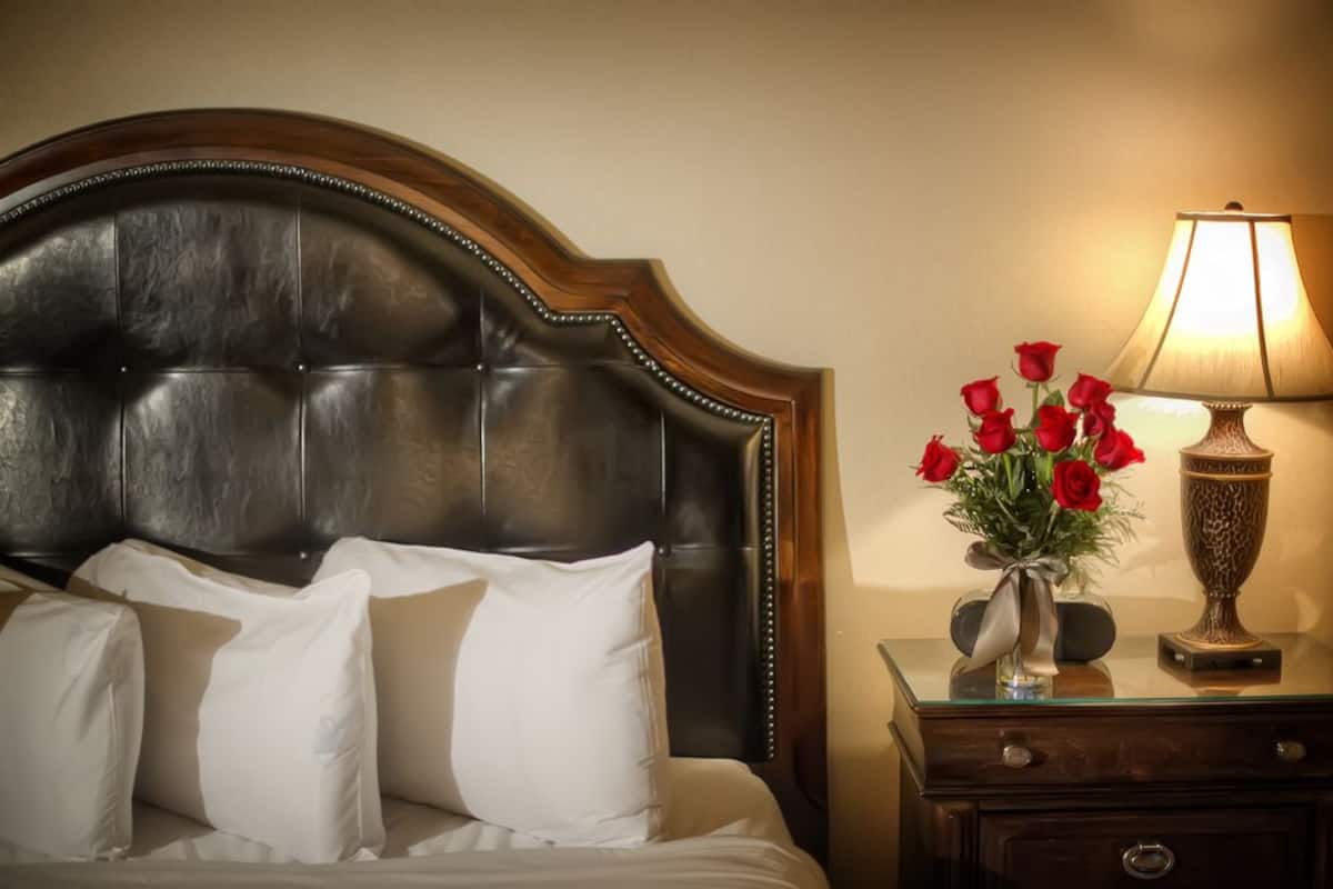 A bed with a dark headboard, white pillows, and a vase with red roses on a bedside table.