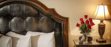 Una cama con cabecera oscura, almohadas blancas y un florero con rosas rojas sobre una mesita de noche.