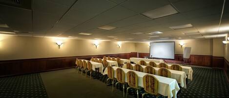 A conference room with rows of tables and chairs, a projector screen, and wall-mounted lighting.