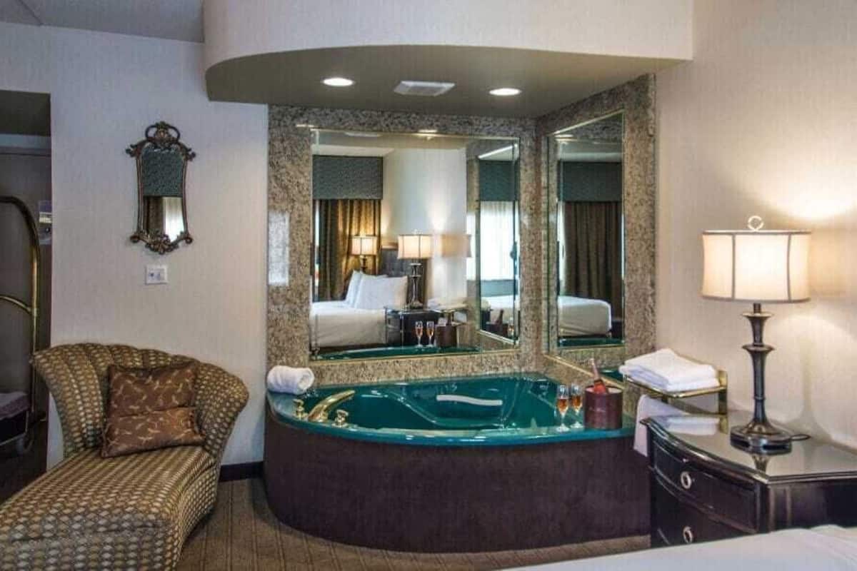 Habitación de hotel con cama curvada, zona de estar con una silla y una mesita, espejo grande, mesita de noche con lámpara y bañera.