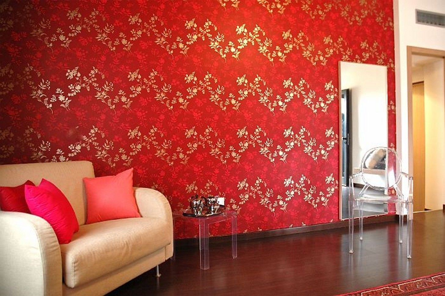 A room with a red floral wallpaper, a beige sofa with red cushions, a clear glass coffee table, and a clear chair.