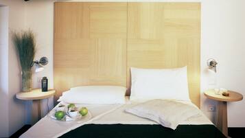 Una cama bien hecha con sábanas blancas, cabecera de madera y mesitas de noche con lámparas y un florero con follaje.