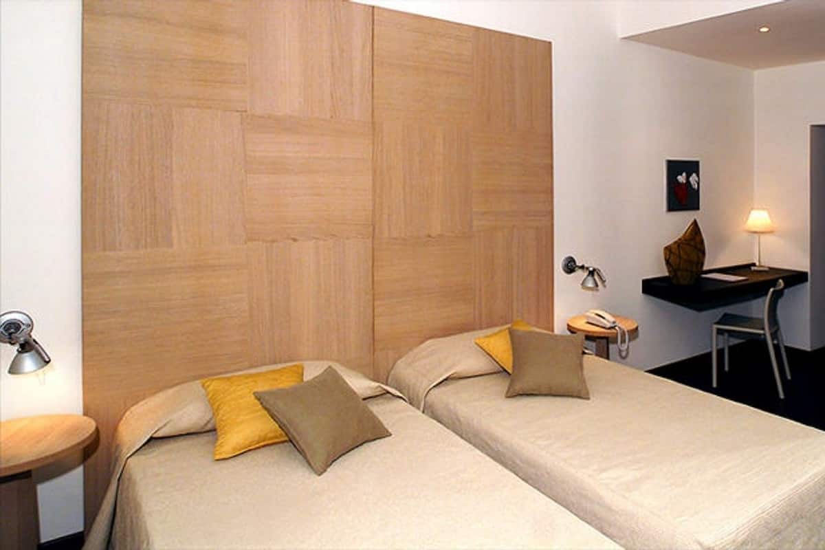 A hotel room with two beds, a desk, a chair, a lamp, and a small table.