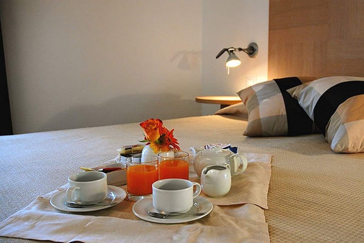 A neatly made bed with a pillow, a bedside table with a lamp, and a breakfast setup including cups, a teapot, and orange juice.