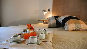 Una cama bien hecha con un almohadón, una mesita de noche con lámpara y una bandeja de desayuno con tazas, tetera y zumo de naranja.