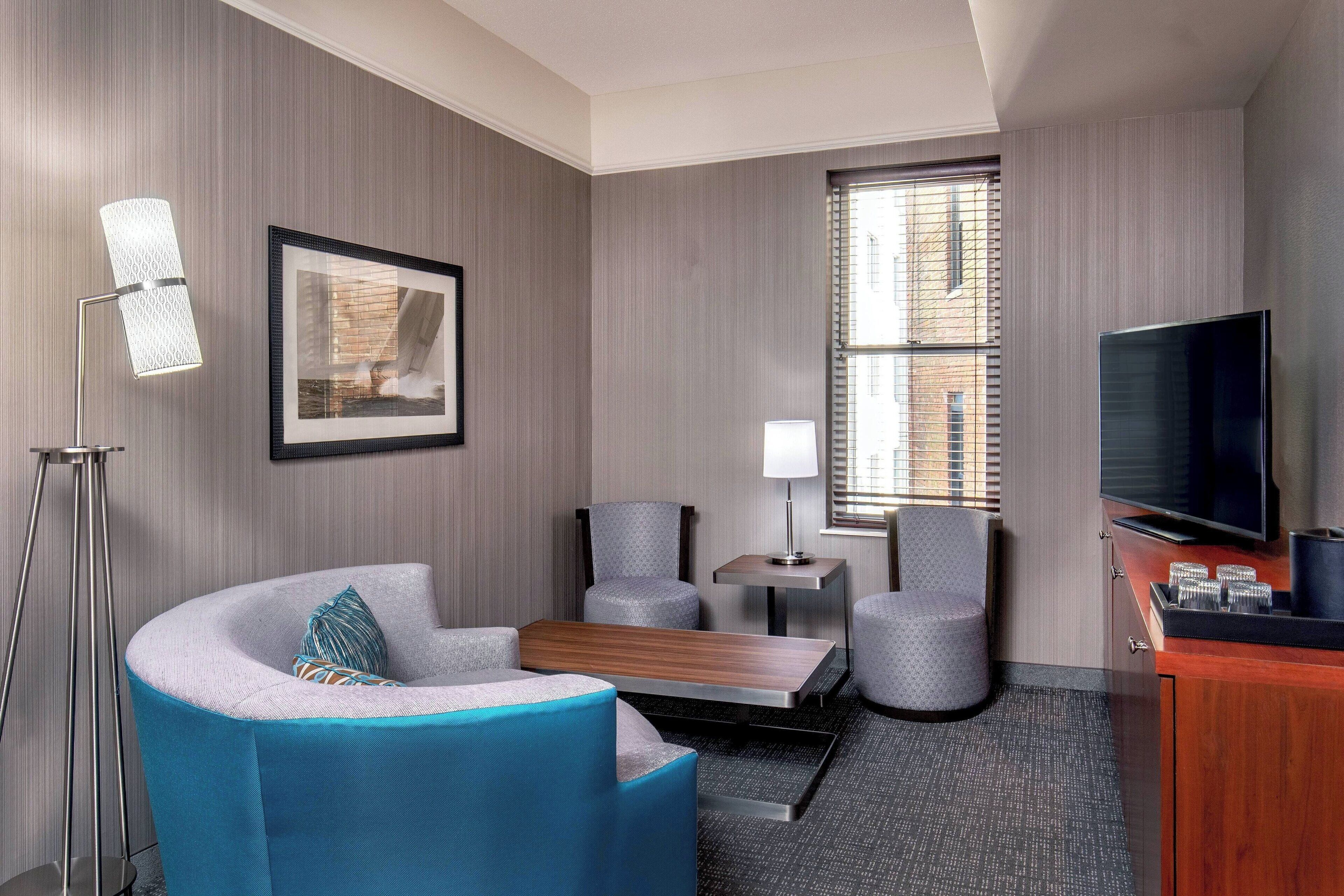 Une chambre d’hôtel avec un canapé incurvé, deux fauteuils, une petite table, un téléviseur et un tableau encadré au mur.
