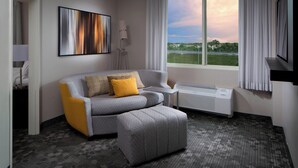 A modern hotel room with a grey sofa, a matching ottoman, a flat-screen TV, a standing lamp, and a large window with a view of a green landscape.