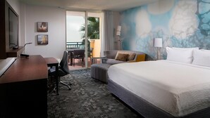 A hotel room with a large bed, a desk with a computer, a chair, a sofa, and a balcony with a view of palm trees.