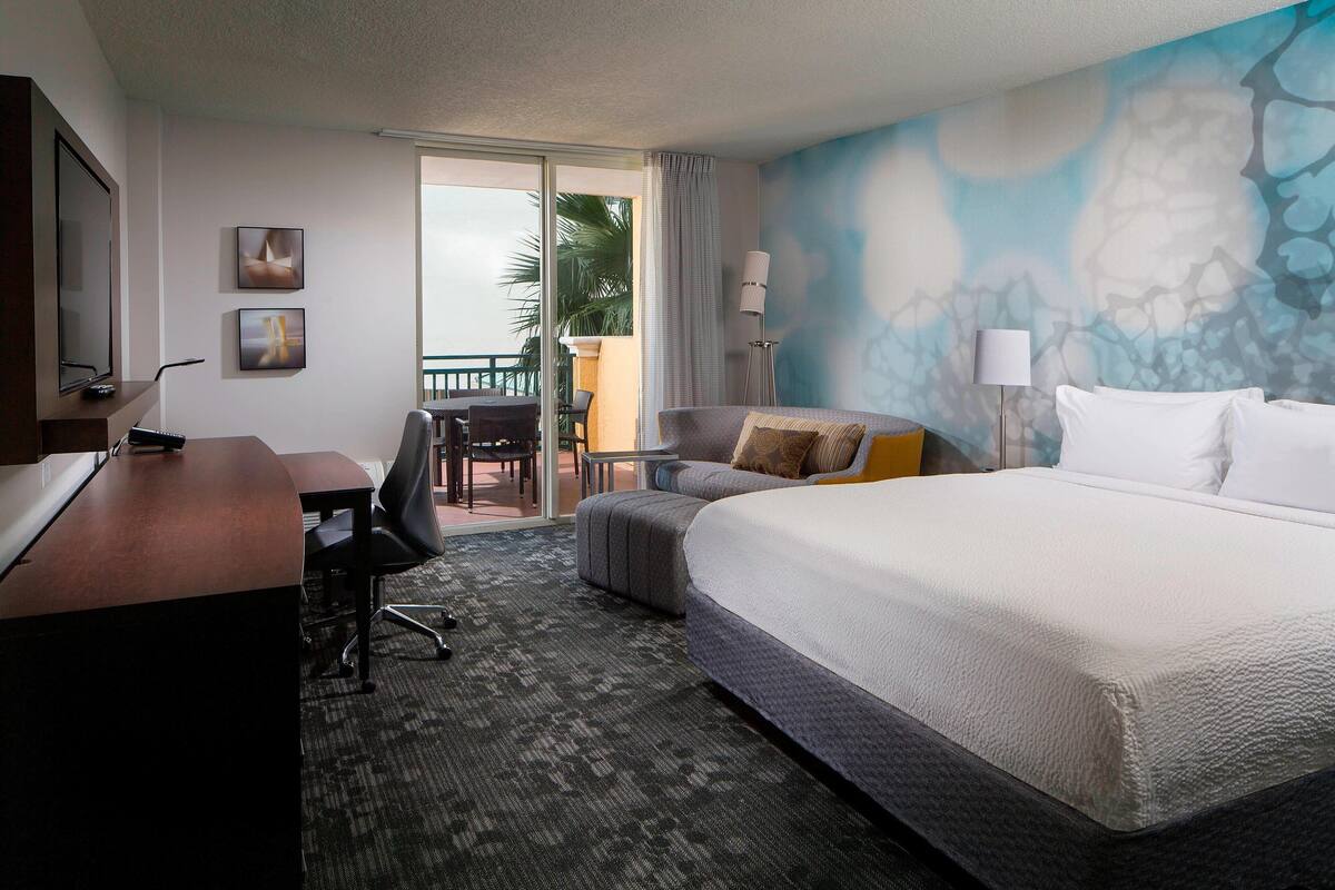 A hotel room with a large bed, a desk with a computer, a chair, a sofa, and a balcony with a view of palm trees.