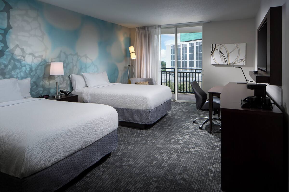 A hotel room with two beds, a desk, a chair, a lamp, and a view of the city through a large window.