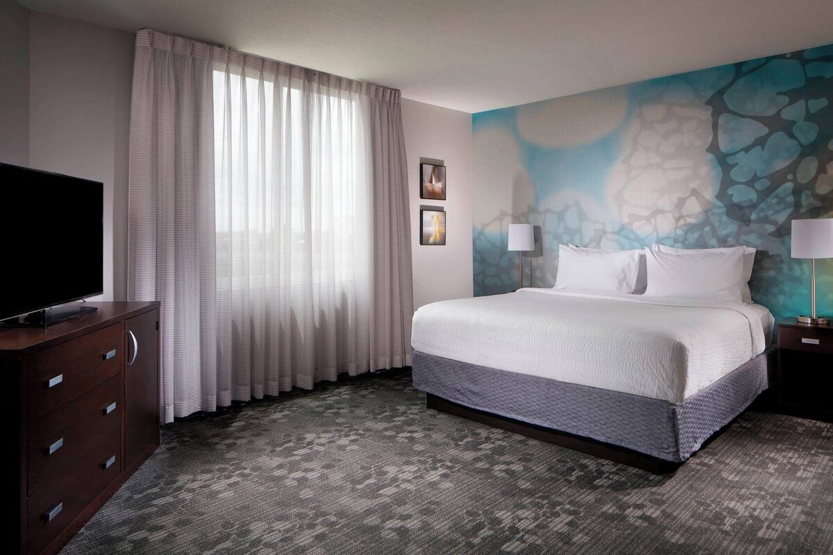 A hotel room with a large bed, a flat-screen TV on a wooden cabinet, a window with sheer curtains, and a wall with a textured mural.