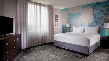 Habitación de hotel con una cama grande, un televisor de pantalla plana sobre un mueble de madera, una ventana con cortinas translúcidas y una pared con un mural texturizado.