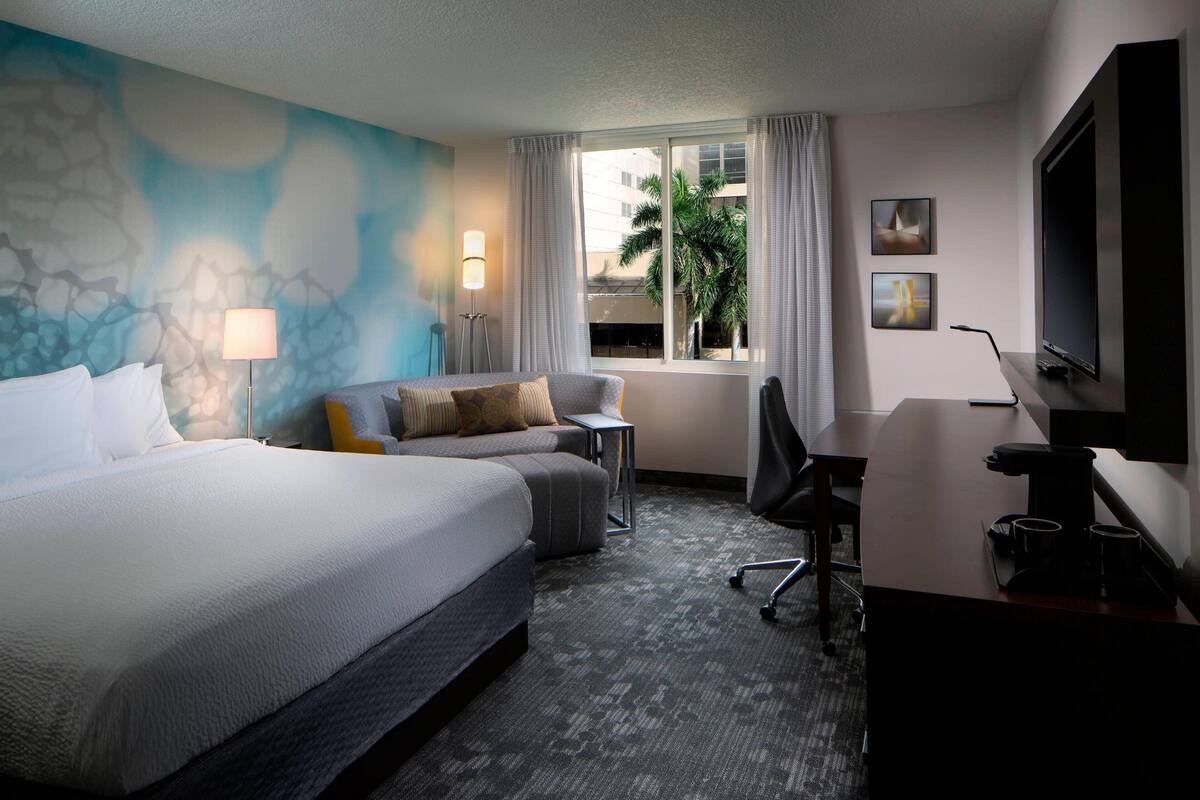 A hotel room with a large bed, a desk, a chair, a TV, and a window with curtains.
