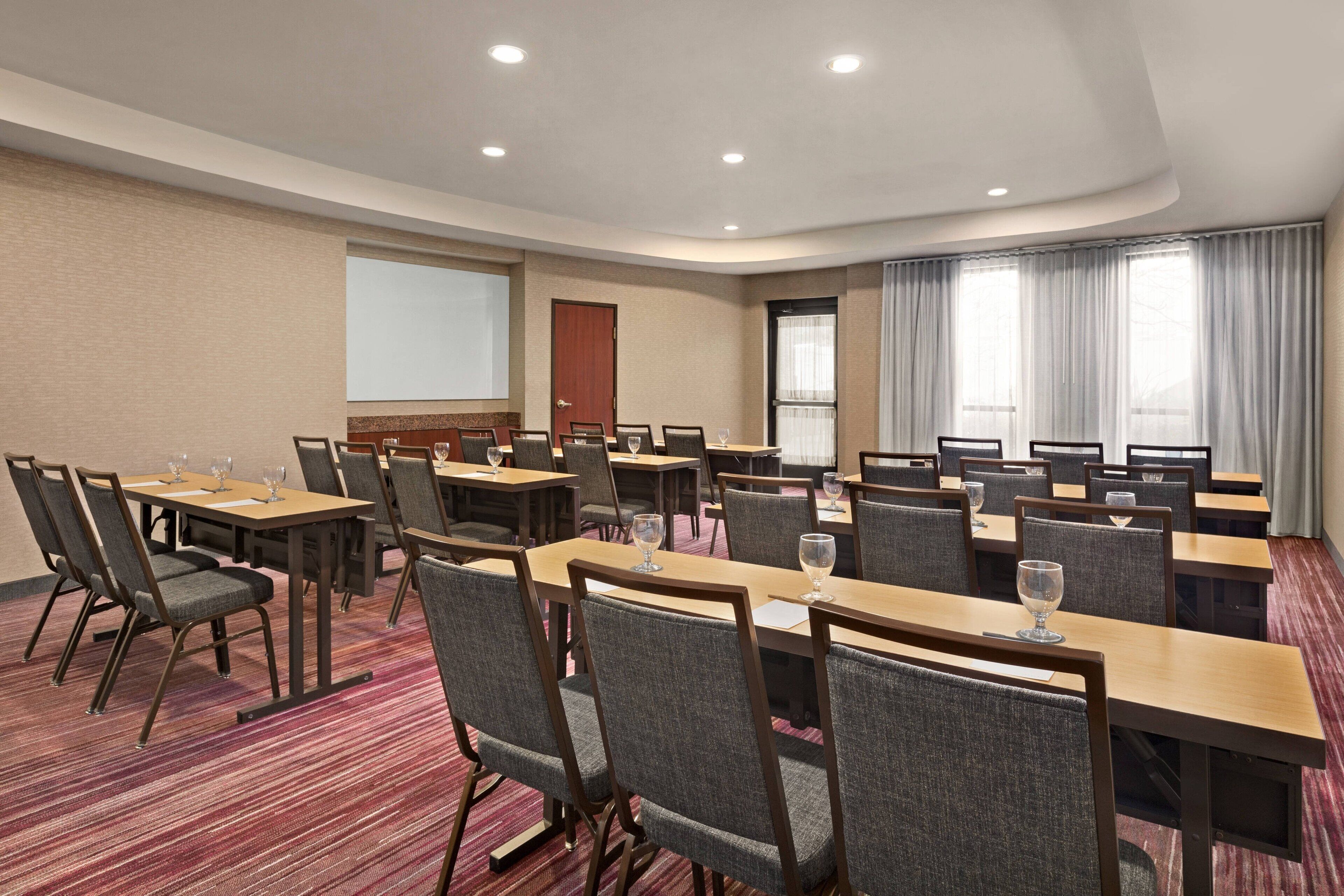A conference room with multiple tables and chairs, a projector screen, and a large window with curtains.