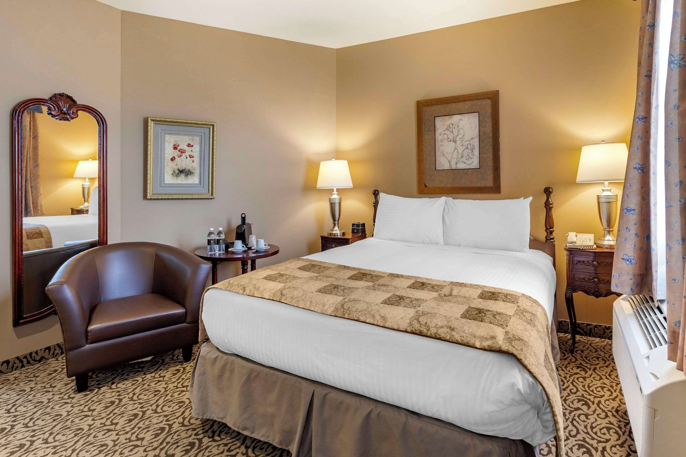 A hotel room with a large bed, a leather armchair, a bedside table with a lamp, and a wall-mounted picture.