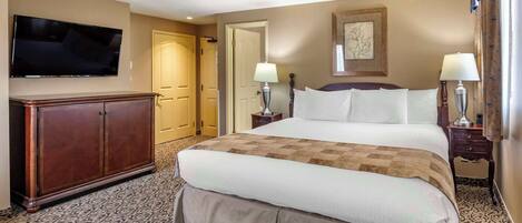 A hotel room with a large bed, a TV mounted on the wall, a wooden dresser, two bedside lamps, and a patterned carpet.