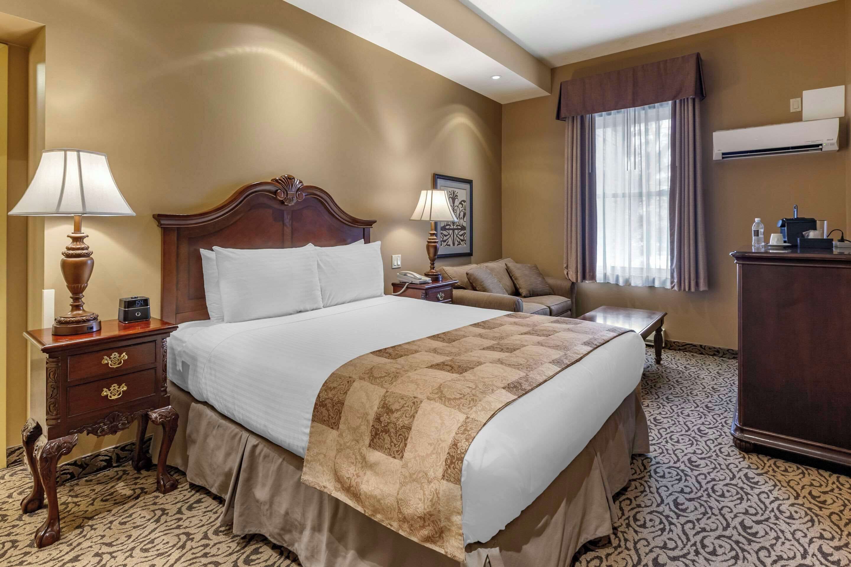 A hotel room with a large bed, a bedside table with a lamp, a small table, a sofa, and an air conditioning unit.