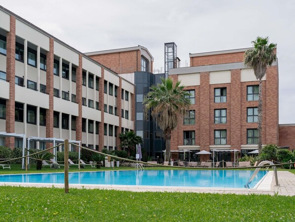 A modern building with a swimming pool, lounge chairs, and palm trees. - Mercure Rome Leonardo da Vinci Airport (Fiumicino)