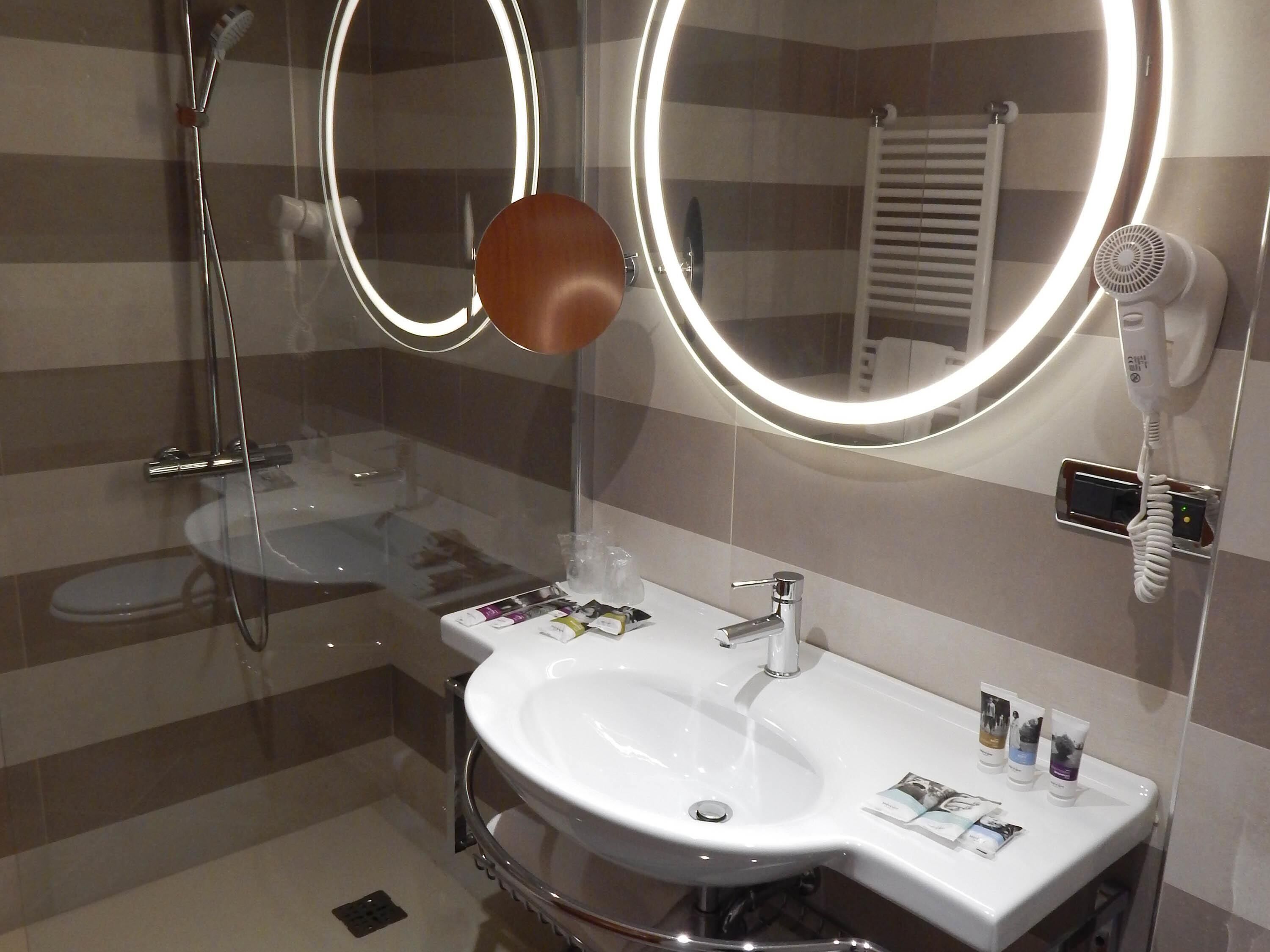 A modern bathroom with a glass-enclosed shower, a white sink with a mirror above it, and a hairdryer mounted on the wall.
