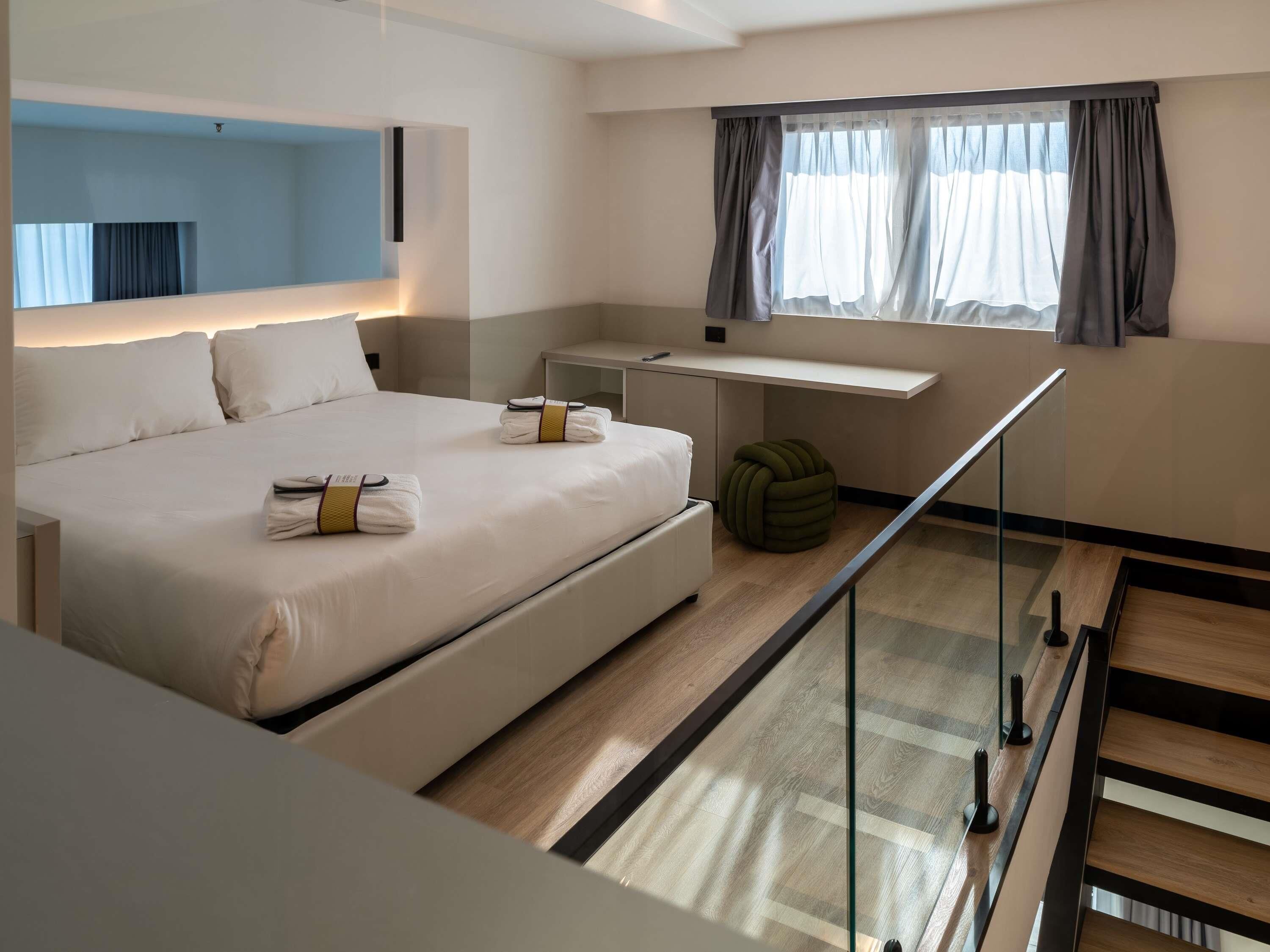 A modern hotel room with a large bed, a desk, and a view of the city through the window.