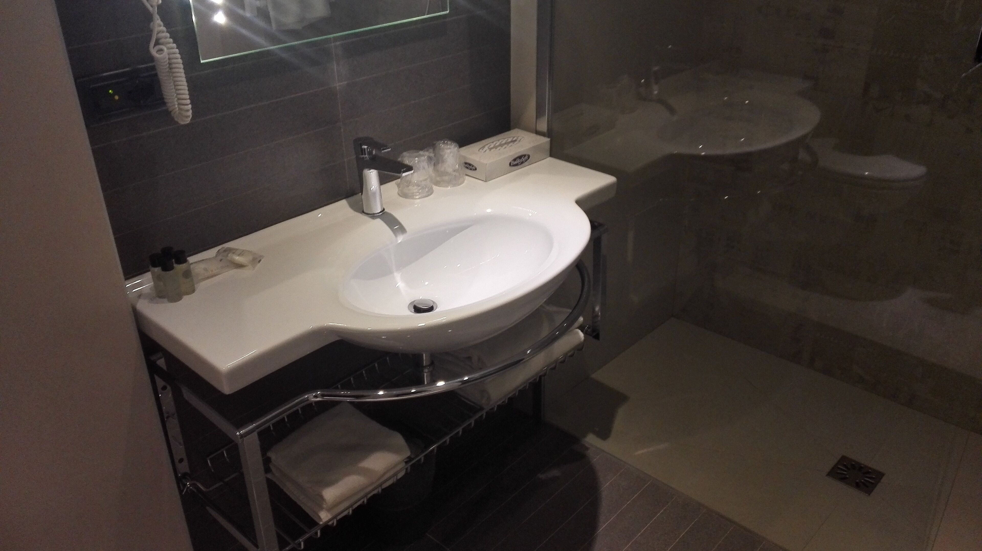 Un baño moderno con un lavamanos blanco, cabina de ducha con mampara de vidrio y pared revestida de baldosas oscuras.