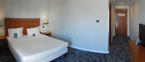 Una habitación de hotel con una cama grande, una mesita de noche con lámpara, un televisor sobre un aparador de madera y suelo cubierto de alfombra.