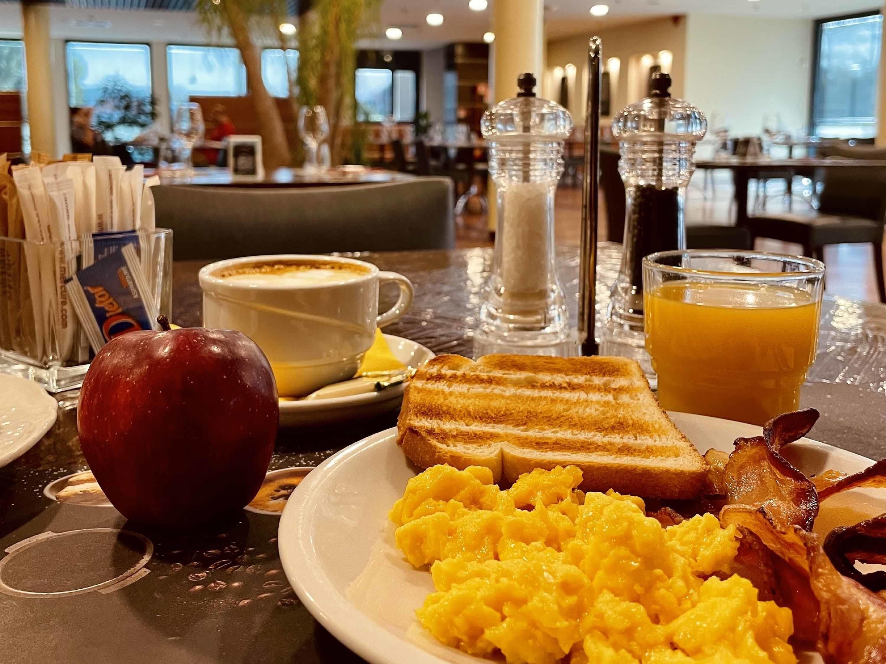 Un desayuno con huevos revueltos, tostadas, tocino, un vaso de jugo de naranja, una taza de café y una manzana roja sobre una mesa de hotel.