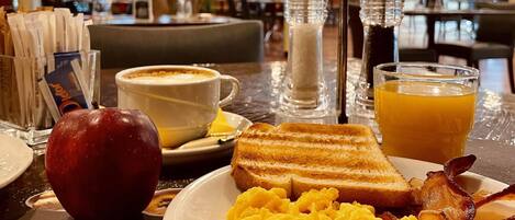 Un desayuno con huevos revueltos, tostadas, tocino, un vaso de jugo de naranja, una taza de café y una manzana roja sobre una mesa de hotel.