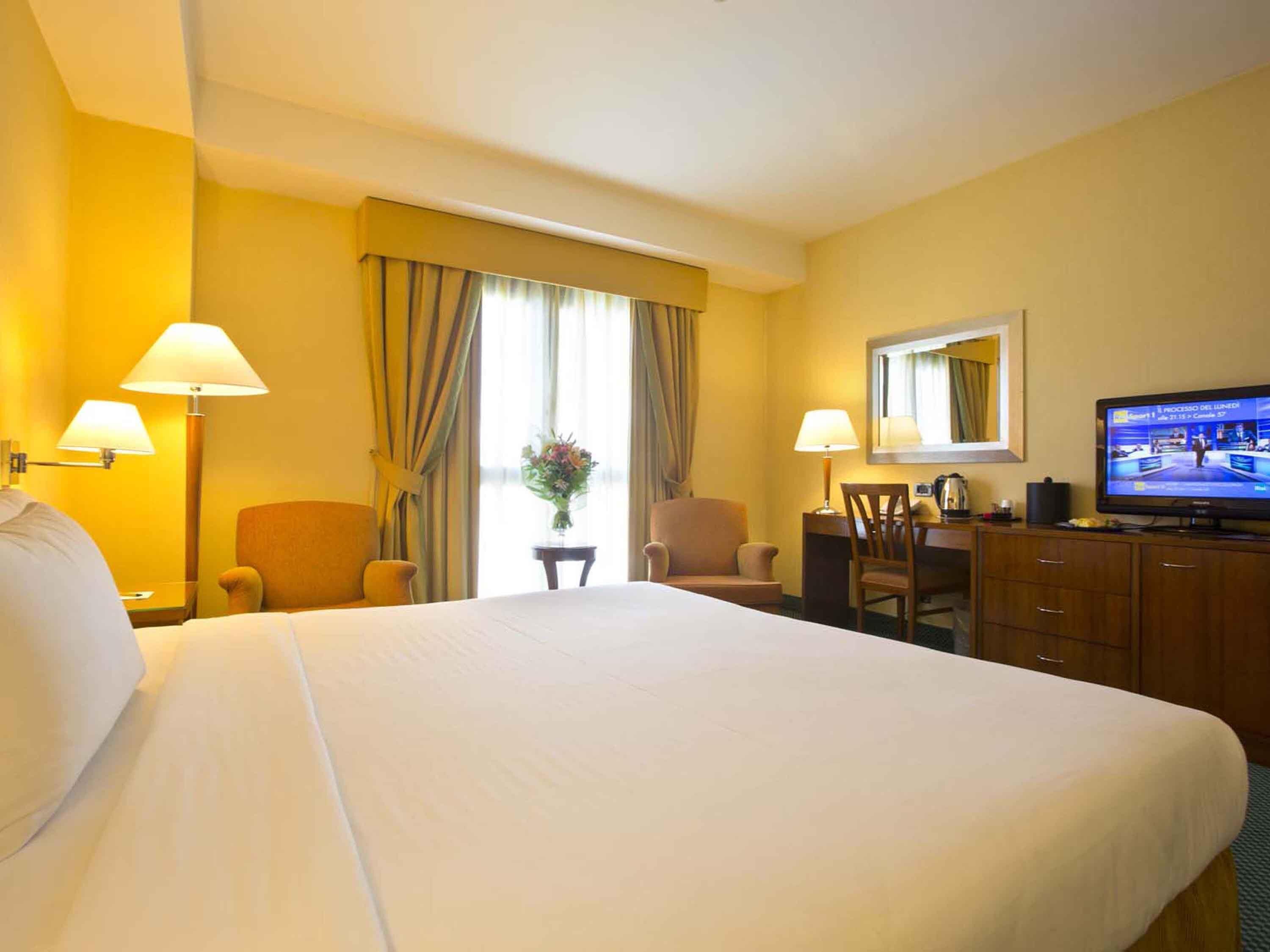 A hotel room with a large bed, two armchairs, a desk with a television, and a window with curtains.