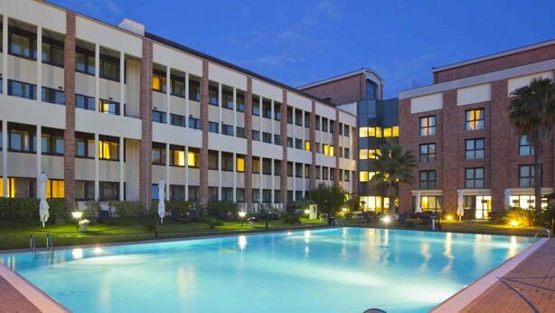 A hotel with a swimming pool, multiple floors, and a well-lit exterior. - Mercure Rome Leonardo da Vinci Airport (Fiumicino)