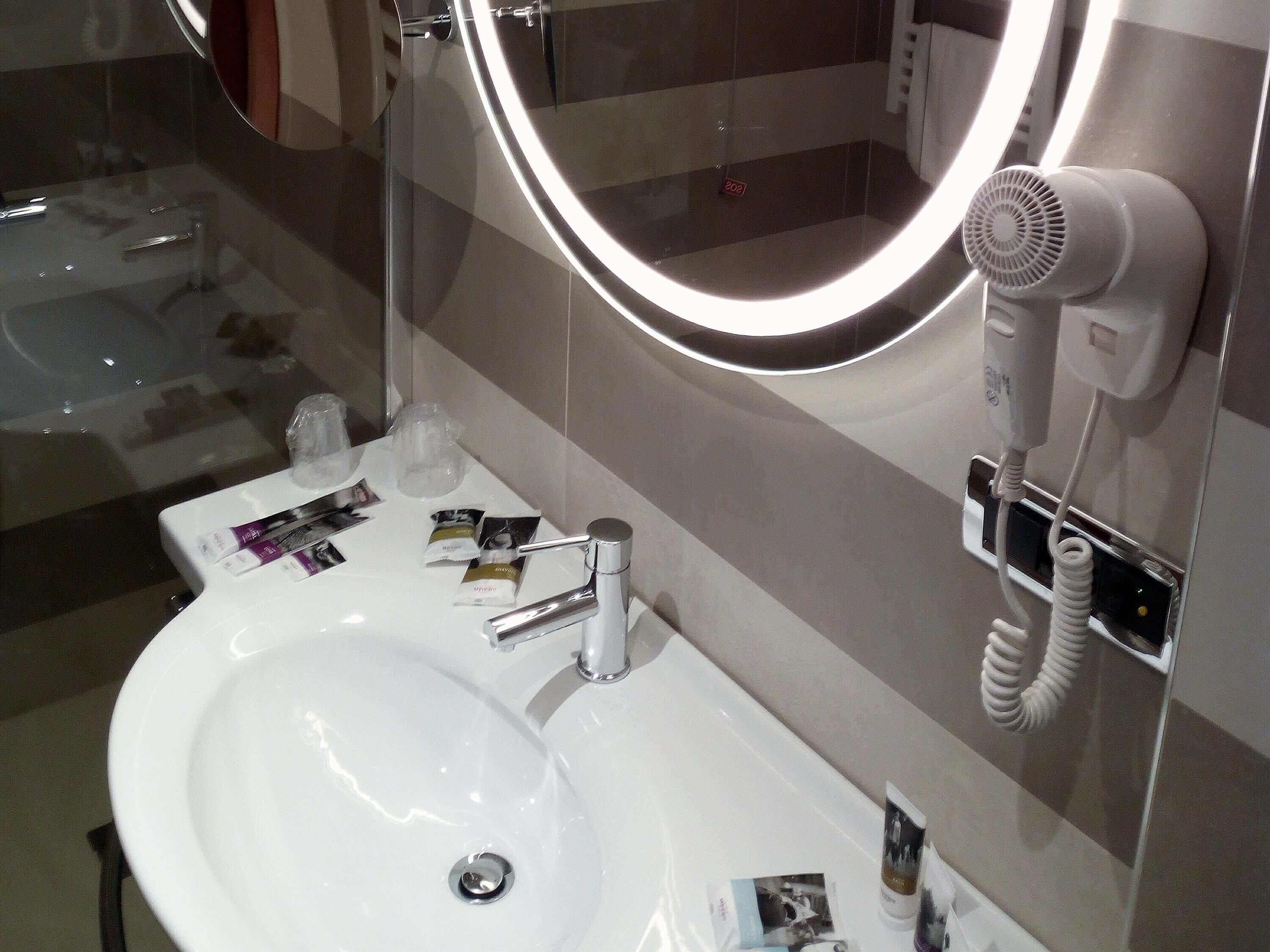 A modern bathroom with a white sink, a mirror with a light, a hairdryer, and toiletries.