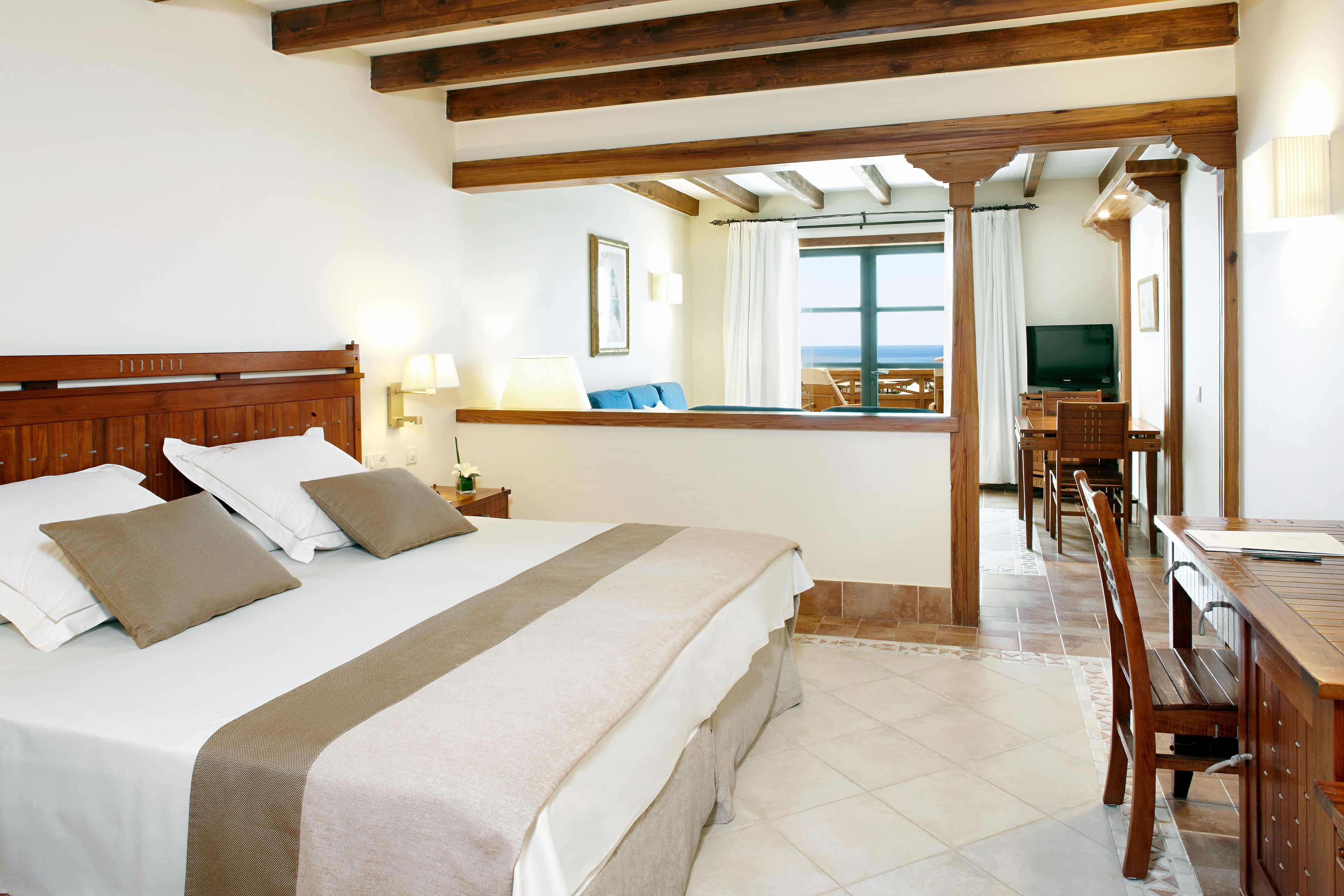 Una habitación de hotel con una cama grande, cabecera de madera, un escritorio con una silla, un televisor y vista al mar a través de una ventana.
