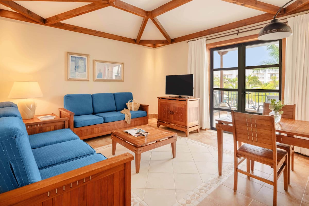 Ein Wohnzimmer mit blauem Sofa, einem Holztisch und einem Fernseher. Ein Balkon bietet Blick auf Palmen.