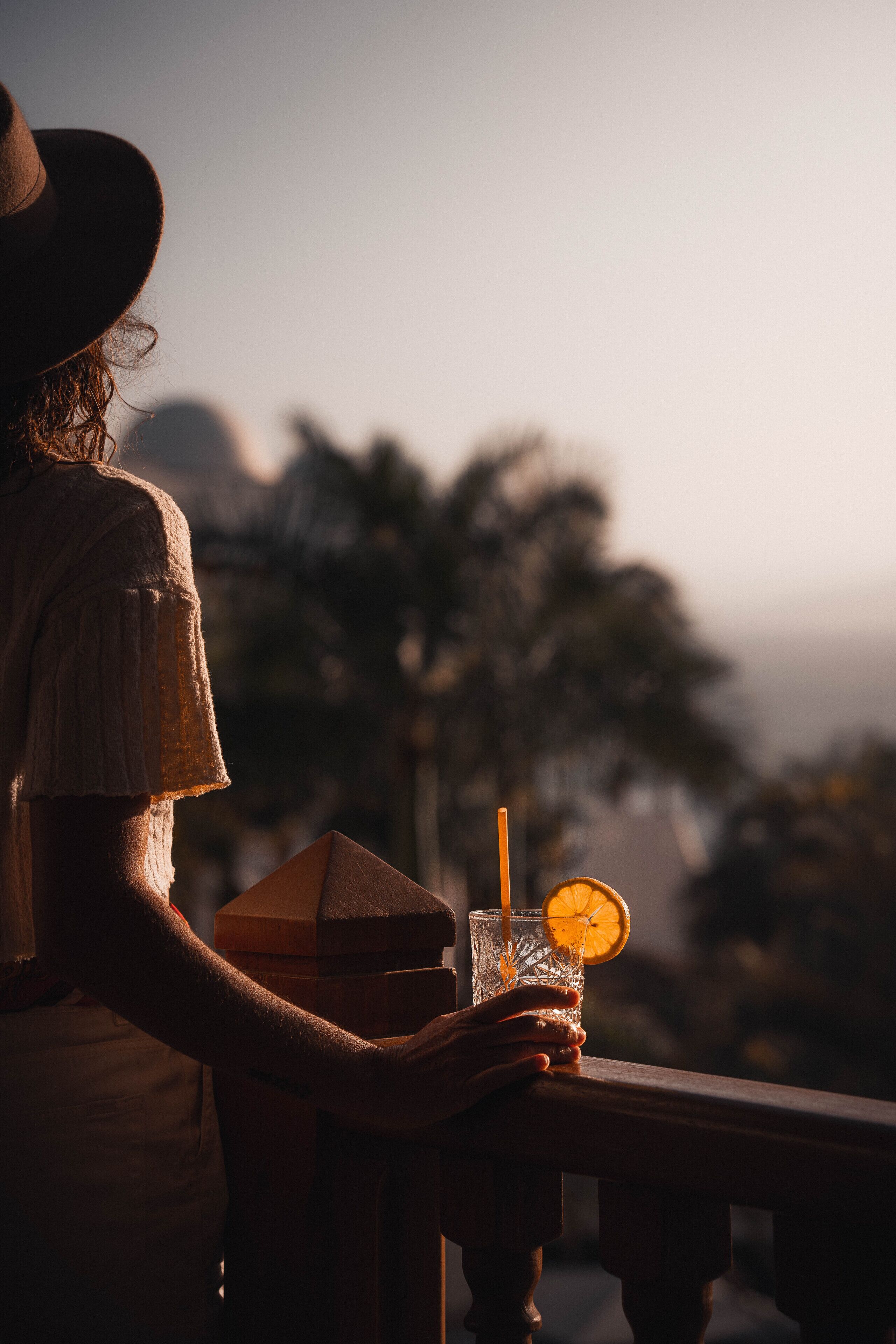 Una persona sostiene una bebida con una rodaja de naranja, sentada en un balcón con vistas a palmeras.