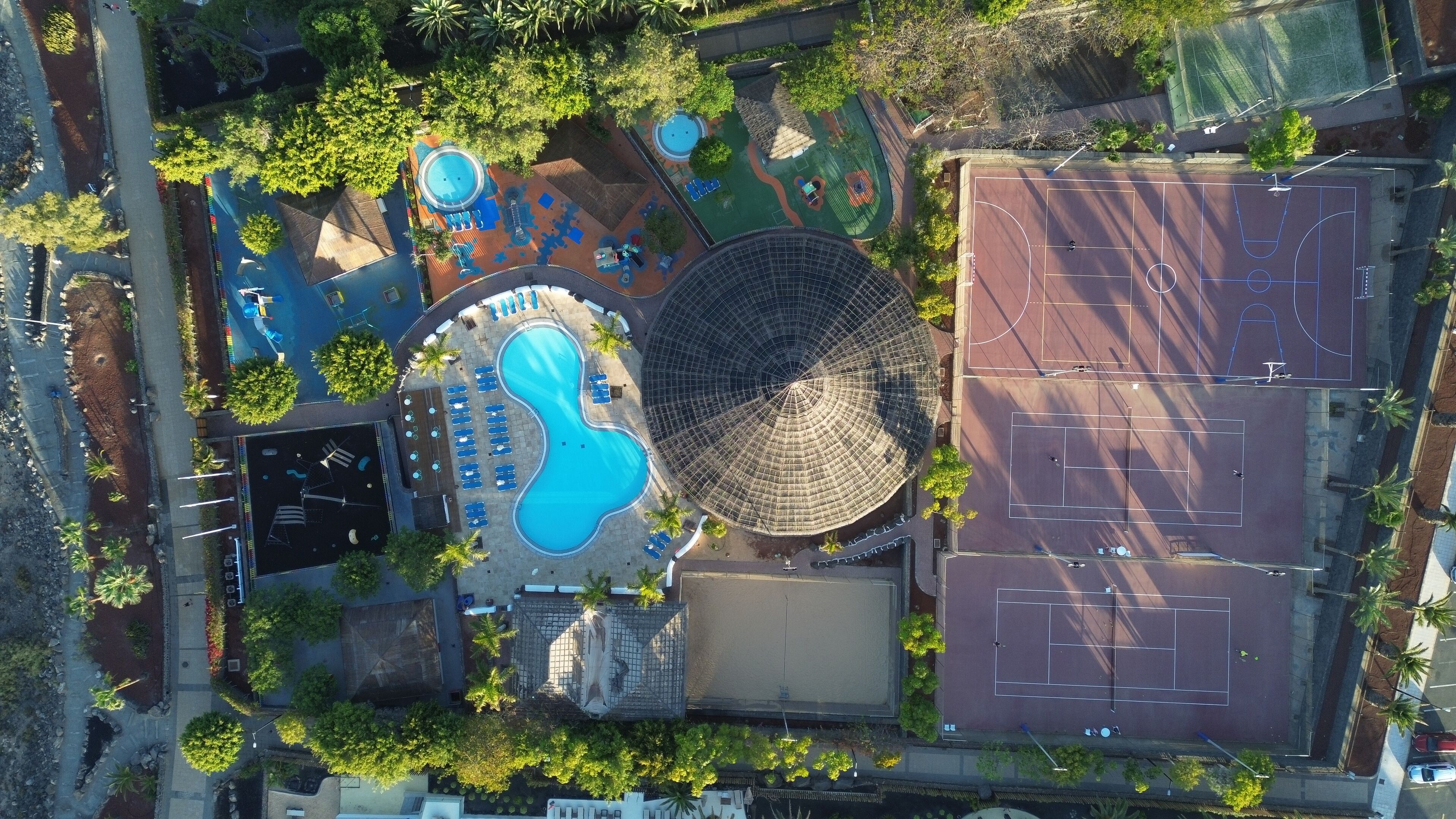Vista aérea de un área recreativa con piscina, canchas de tenis y zona de estar techada.