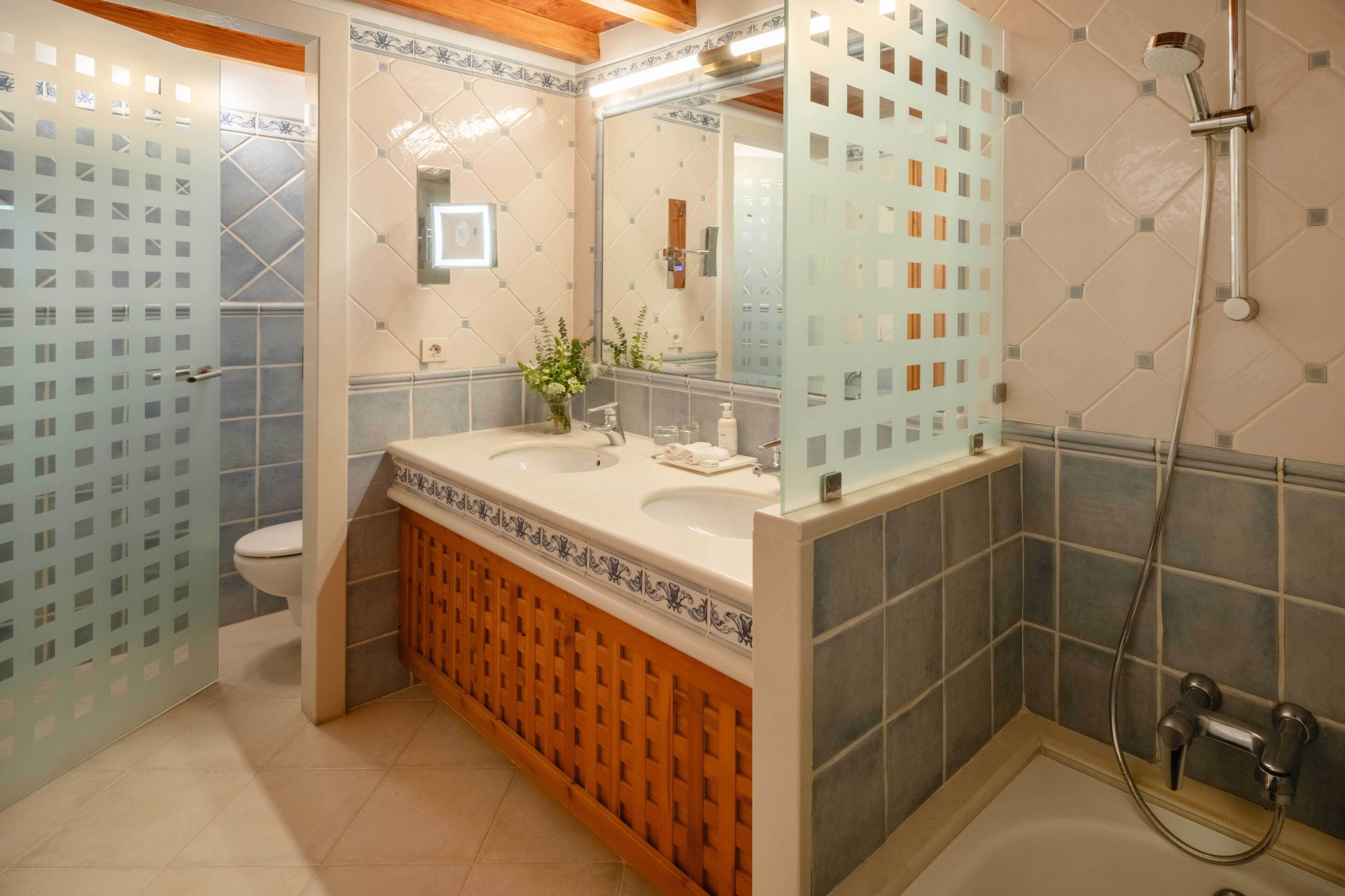 Ein Badezimmer mit einem Doppelwaschtisch, Toilette, Duschbereich und Spiegel mit dekorativer Fliesenfassade.
