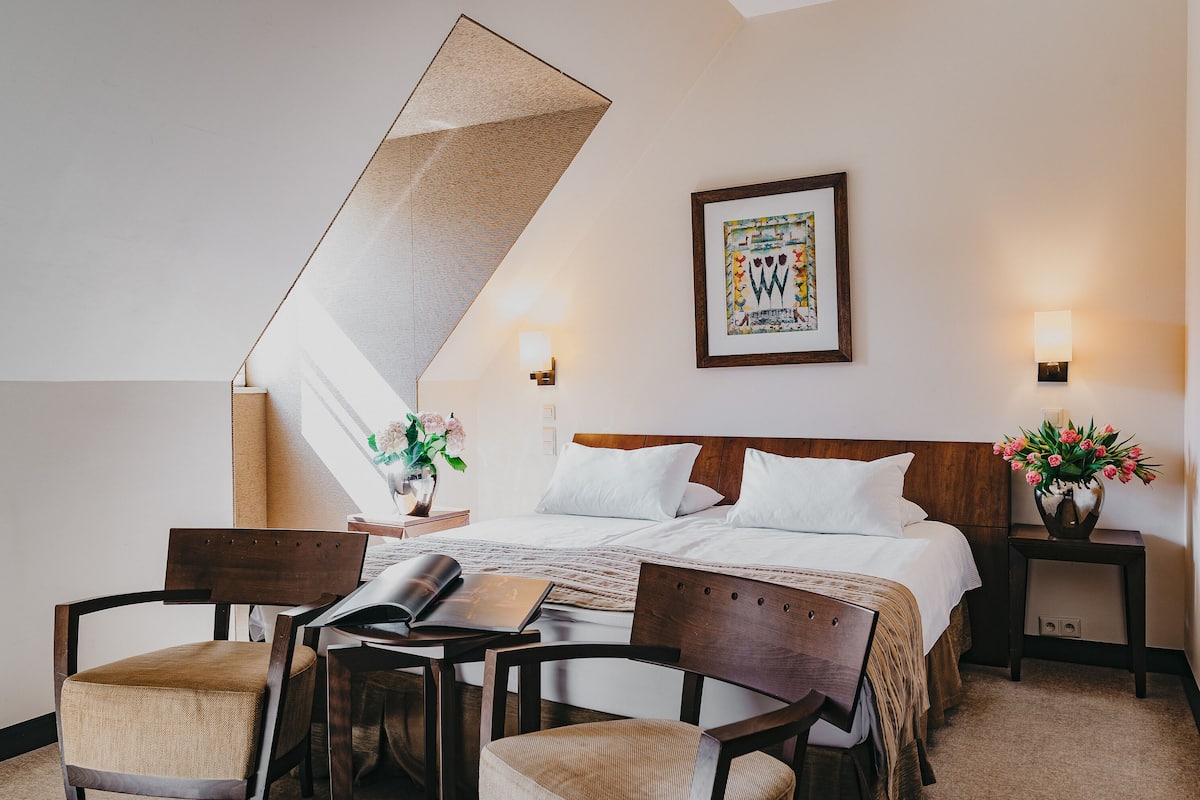 Une chambre d’hôtel comprenant un lit, un bureau, deux chaises, une table de chevet avec un vase de fleurs, une lampe fixée au mur et un tableau encadré.