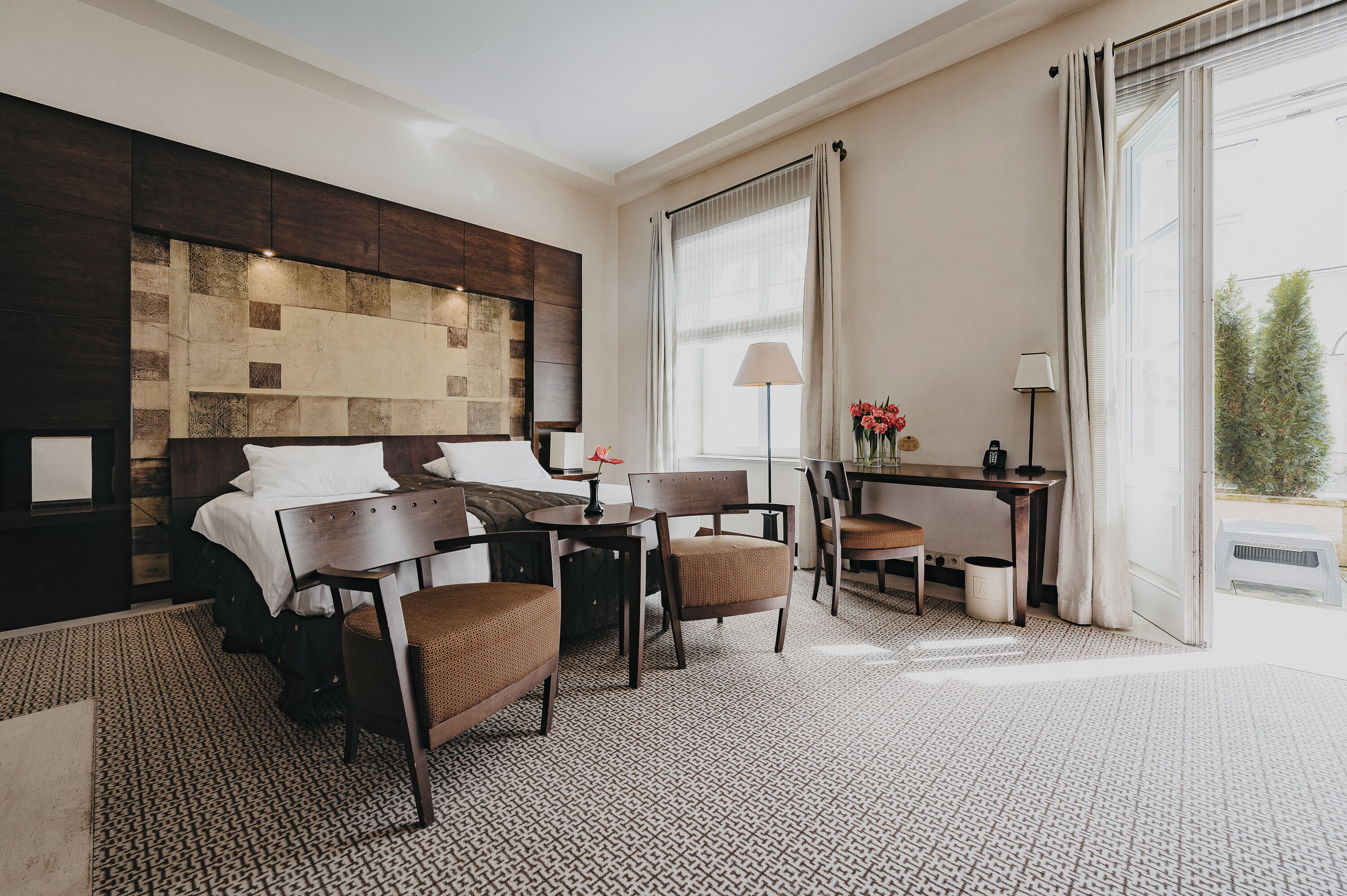 A hotel room with a bed, a desk, two chairs, a lamp, and a view of the outdoors.