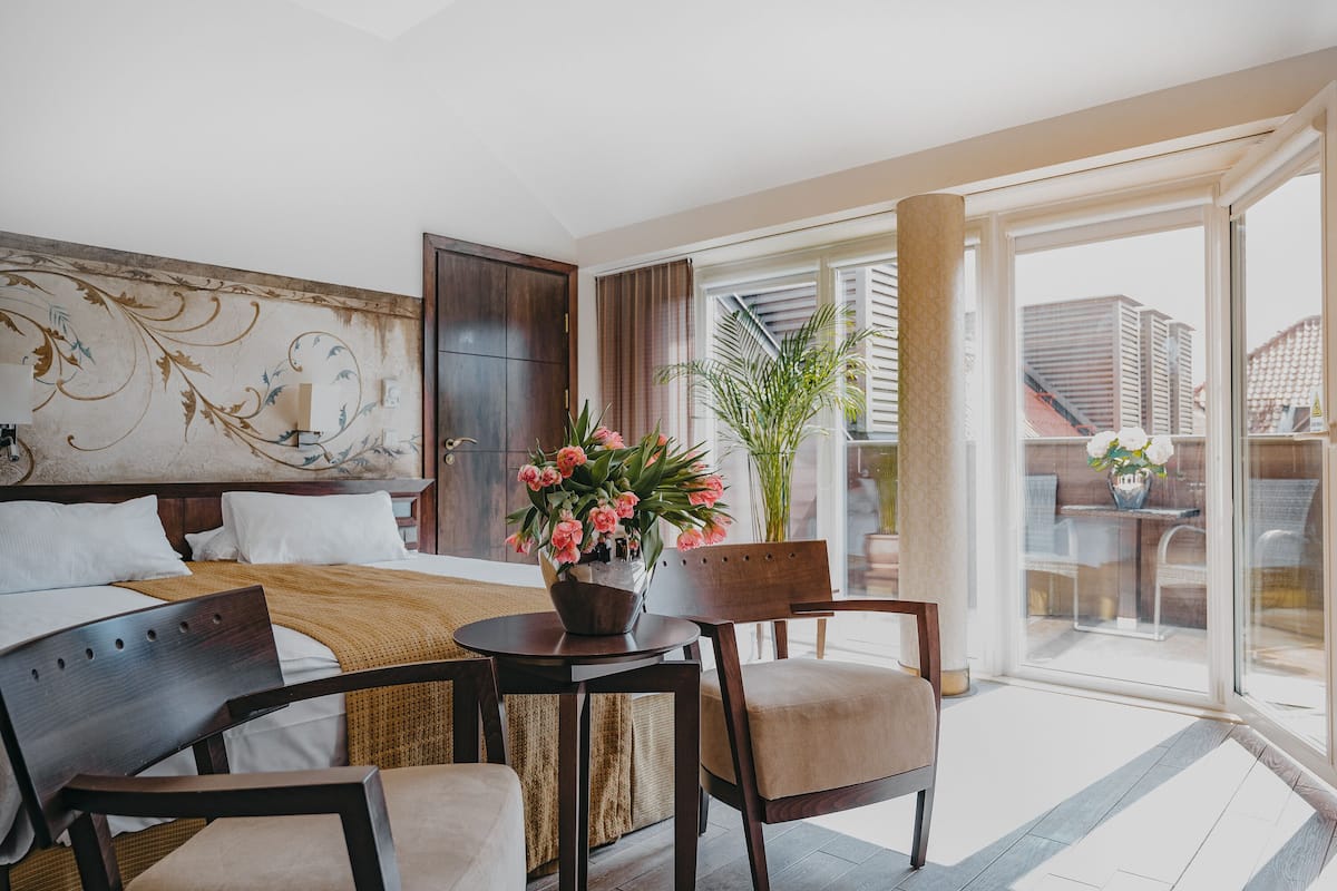 Une chambre d’hôtel avec un lit, une table à manger avec des chaises, une grande fenêtre donnant sur des immeubles et une plante en pot avec des fleurs.