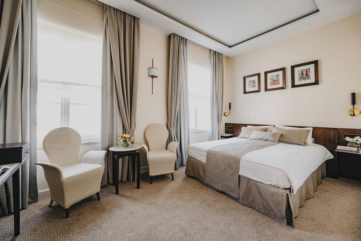 Une chambre d’hôtel avec un lit, deux chaises, une petite table sur laquelle est posé un vase de fleurs et une table de chevet avec une lampe.