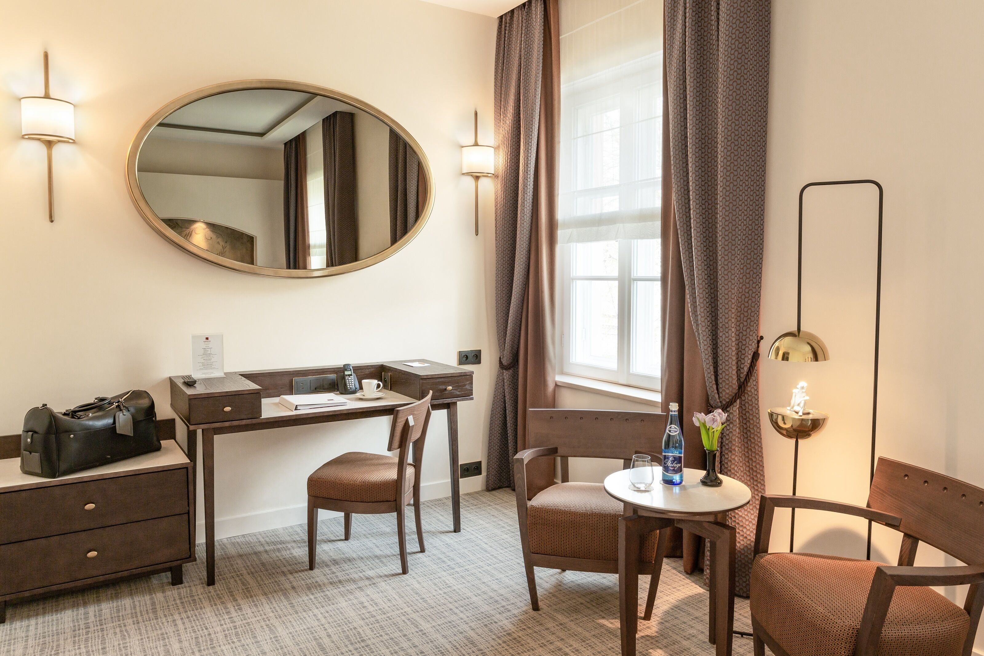A hotel room with a desk, two chairs, a small table with a bottle and flowers, a mirror, and a lamp.