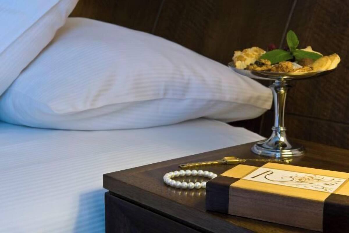 Un lit bien fait, avec du linge de lit blanc, une table de chevet sur laquelle se trouve un plateau décoratif contenant un dessert, un bracelet de perles et une carte avec un monogramme.